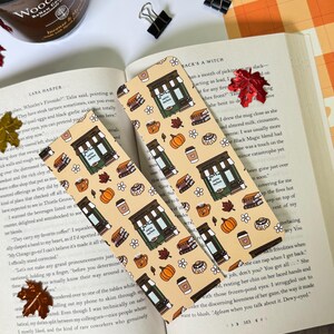 Könnte beinhalten: Zwei beige Lesezeichen mit einem sich wiederholenden Muster aus einem Café, Kaffeetassen, Kürbissen und Büchern. Die Lesezeichen liegen auf einem offenen Buch. Herbstliche Accessoires sind darum herum angeordnet.