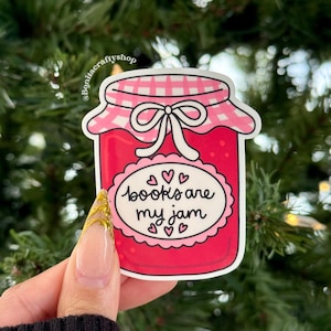 May include: A sticker featuring a jar of jam with a pink lid and a white bow. The jar is filled with red jam and has a label that reads "books are my jam" with hearts. The sticker is held against a background of green foliage.