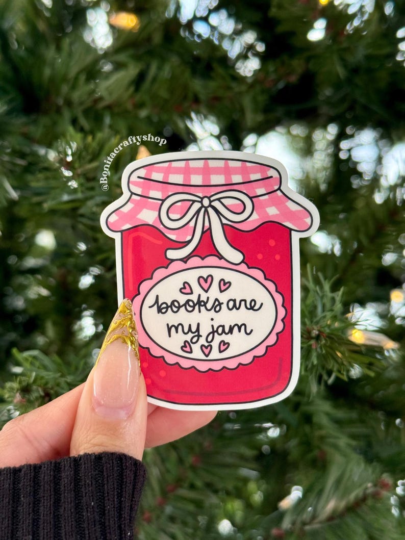May include: A sticker featuring a jar of jam with a pink lid and a white bow. The jar is filled with red jam and has a label that reads "books are my jam" with hearts. The sticker is held against a background of green foliage.