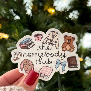 May include: A sticker with the text "the homebody club" and illustrations of a mug, books, a sweater, a teddy bear, socks, a bunny, a blanket, a bow, and a book. The sticker is held by a hand with red nail polish.