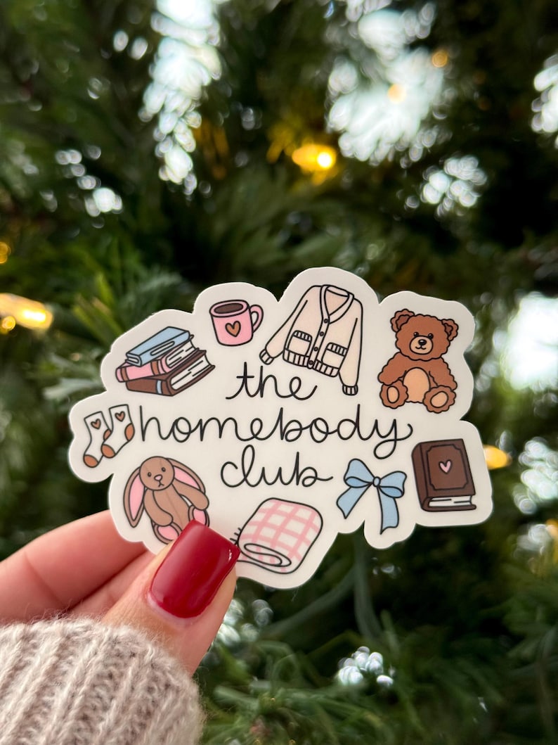 May include: A sticker with the text "the homebody club" and illustrations of a mug, books, a sweater, a teddy bear, socks, a bunny, a blanket, a bow, and a book. The sticker is held by a hand with red nail polish.