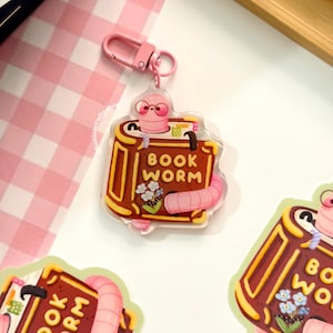 May include: A clear acrylic keychain featuring a brown book with the text BOOK WORM printed on the cover. A pink worm wearing glasses emerges from the top of the book, and a small cluster of blue flowers is illustrated on the front cover.