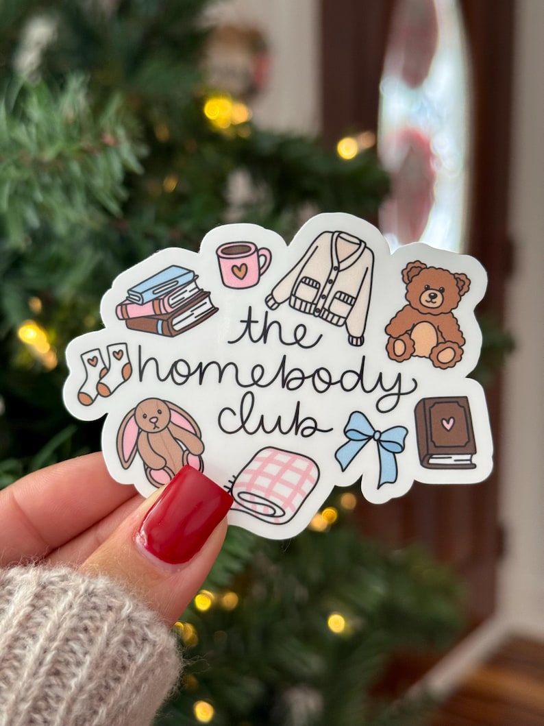May include: A sticker with the text "the homebody club" and illustrations of a mug, books, a sweater, a teddy bear, socks, a bunny, a blanket, a bow, and a book. The sticker is held by a person with red nail polish.