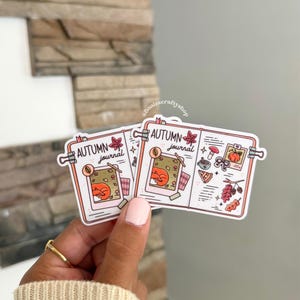 May include: Two die-cut vinyl stickers featuring an open journal design with the text "AUTUMN journal". The illustrations include a fox, a pumpkin, a mushroom, a slice of pie, and autumn leaves in shades of orange, red, and brown on a white background.