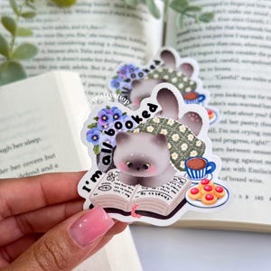May include: A sticker featuring a cartoon cat reading a book, with the text "I'm all booked." The cat is light gray with big eyes, wearing a floral scarf. A cup of coffee and cookies are also depicted. The sticker has a whimsical, bookish theme.