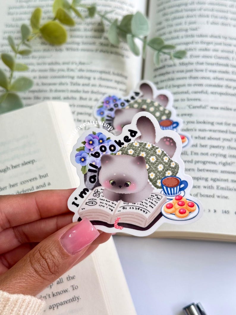 May include: A sticker featuring a cartoon cat reading a book, with the text "I'm all booked." The cat is light gray with big eyes, wearing a floral scarf. A cup of coffee and cookies are also depicted. The sticker has a whimsical, bookish theme.