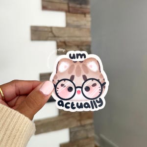 May include: A die-cut sticker featuring a cartoon cat with brown tabby markings wearing large black-rimmed glasses. The text 'um' is printed in black above the cat's head, and the word 'actually' is printed in black below the cat's chin.