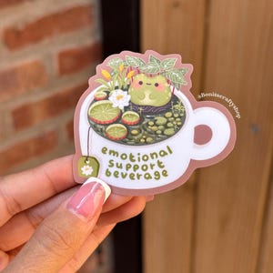 May include: A sticker featuring a white teacup with a frog in a tea-filled cup. The tea includes lime slices, flowers, and leaves. The text "emotional support beverage" is printed on the cup. The sticker is held by a person.