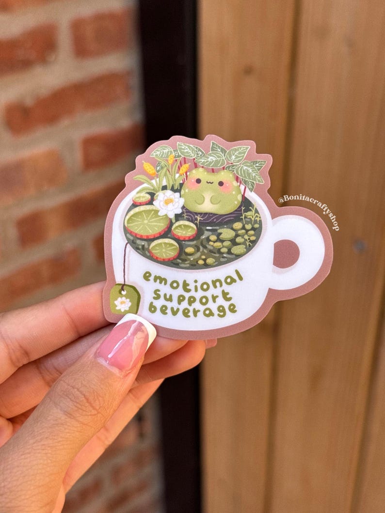 May include: A sticker featuring a white teacup with a frog in a tea-filled cup. The tea includes lime slices, flowers, and leaves. The text "emotional support beverage" is printed on the cup. The sticker is held by a person.