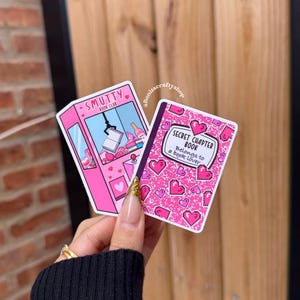 May include: Two stickers are held in a hand. One is pink with the text "Smutty Book Club" and a claw machine graphic. The other is a pink book with hearts and the text "Secret Chapter Book Belongs to a Book Lover".