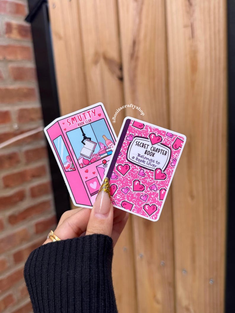 May include: Two stickers are held in a hand. One is pink with the text "Smutty Book Club" and a claw machine graphic. The other is a pink book with hearts and the text "Secret Chapter Book Belongs to a Book Lover".