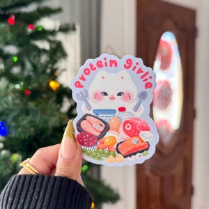 Może przedstawiać: Naklejka z postacią kota z kresk&oacute;wki i napisem "protein girlie". Projekt zawiera obrazy bogatych w białko produkt&oacute;w spożywczych, takich jak łosoś, jajka i jogurt. Naklejka ma kolorowy design.
