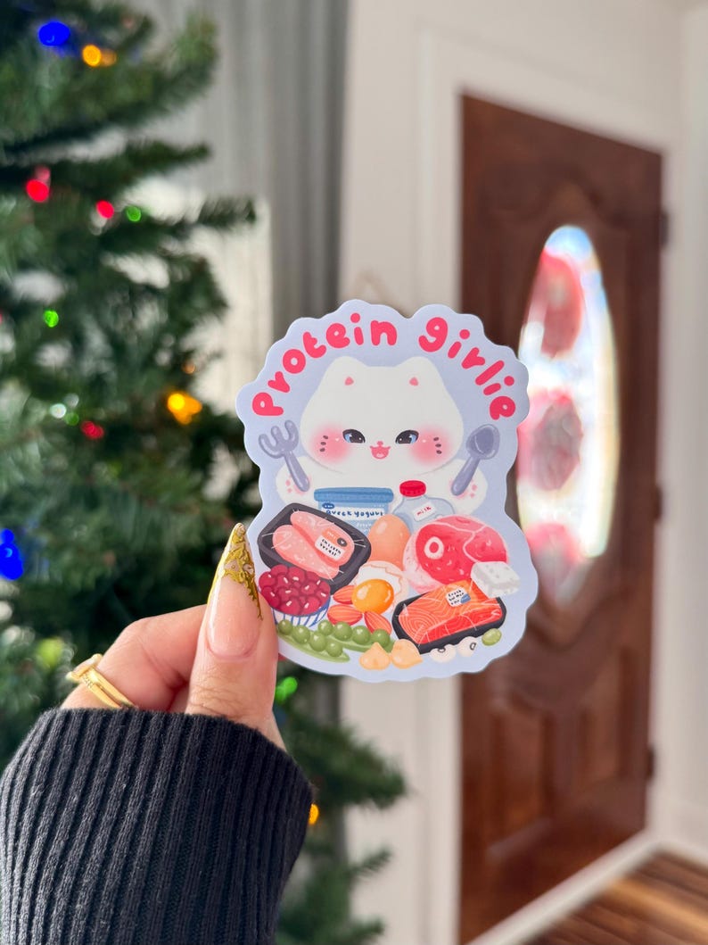 Może przedstawiać: Naklejka z postacią kota z kresk&oacute;wki i napisem "protein girlie". Projekt zawiera obrazy bogatych w białko produkt&oacute;w spożywczych, takich jak łosoś, jajka i jogurt. Naklejka ma kolorowy design.