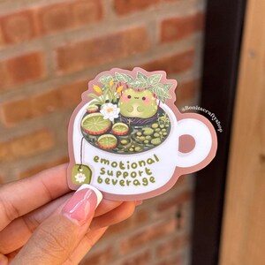 May include: A white sticker in the shape of a teacup with the text "emotional support beverage." The teacup contains a frog, flowers, and tea. A green tea bag with a white flower is attached to the cup.