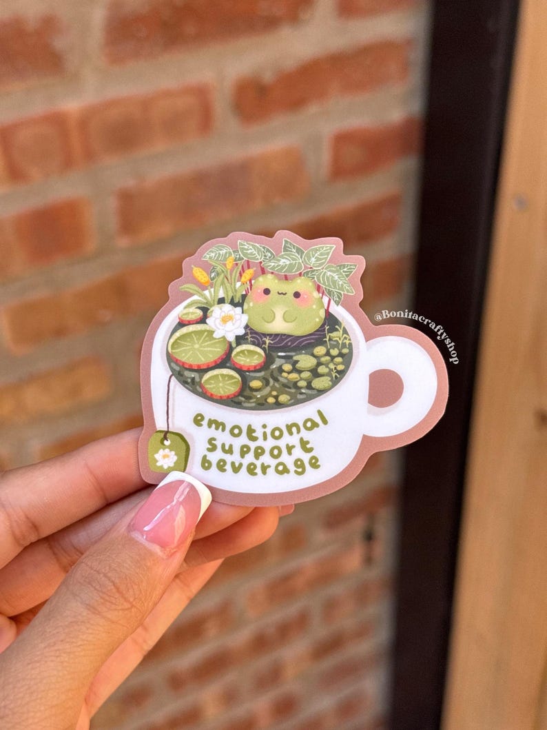 May include: A white sticker in the shape of a teacup with the text "emotional support beverage." The teacup contains a frog, flowers, and tea. A green tea bag with a white flower is attached to the cup.