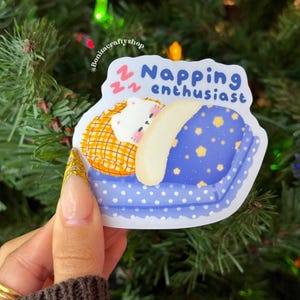 May include: A sticker with a cartoon illustration of a sleeping bear on a blue pillow with yellow stars. The sticker has the text "Napping enthusiast" in blue. The background is a Christmas tree.