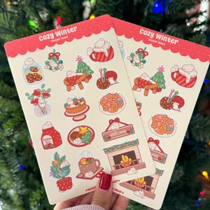 May include: Two sticker sheets with a red scalloped border and the text "Cozy Winter." Each sheet features winter-themed illustrations, including gingerbread cookies, a fireplace, and a mug of hot cocoa. The text "Festive Mind" and "Welcome" are also visible.