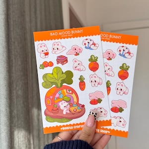 May include: Two sticker sheets with a cute bunny theme. The stickers feature bunnies, carrots, and a bunny house. The text "BAD MOOD BUNNY" is printed on the top of the orange bordered sticker sheets. The stickers are in a variety of poses and colors.