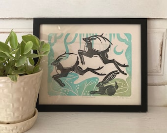 May include: A framed print of a black and white illustration of three animals running through a forest. The animals are a deer and two rabbits. The background is a light blue and green color with a crescent moon in the upper right corner. The print is titled "Wonderlust" and "Into the Hollow".