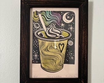 May include: Framed art print of a cosmic matcha drink. The print features a yellow cup with a heart, ice cubes, and whipped cream, set against a starry night sky with a moon and planets. The frame is dark brown.