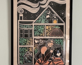 May include: A framed art print featuring a house illustration with a woman and a black cat. The artwork is in black, white, green, and orange. The house has a chimney, windows, and a dress. The background is a starry night.