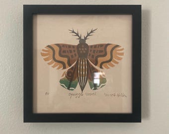 May include: A framed artwork featuring a stylized moth illustration. The moth has brown, orange, and green details with a patterned design. The artwork is in a black frame and is mounted on a light-colored background. The text "Squiggle Moth" and "Nicole Hollow" are visible.