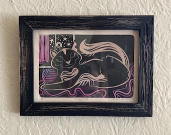 May include: A black and pink linocut print of a cat curled up with a ball of yarn. The print is framed in a black wood frame. The print is signed "Windfall" and numbered "2/2".