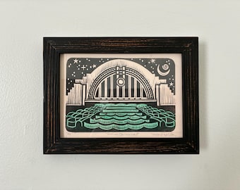 May include: Framed black and white art print with a dark brown wooden frame. The print depicts an archway with a night sky background, a crescent moon, and stars. The bottom of the print features a teal-colored water design.