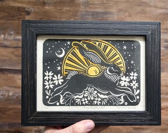 May include: A black framed linocut print featuring a black rabbit leaping through white flowers against a yellow sunburst and starry night sky. The print includes the handwritten text "EV", "Playrabbit III", and a signature at the bottom edge.