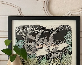 May include: Black framed art print featuring a nighttime landscape with a crescent moon, stars, and a figure. The print has a black and white design with accents of light green and blue. A small, cream-colored vase with green leaves is in the foreground.