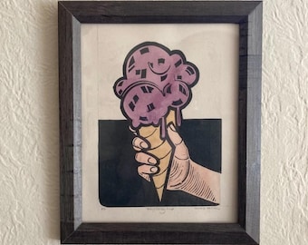 May include: A framed print of a hand holding a melting purple ice cream cone. The print is in a black frame with a white mat.