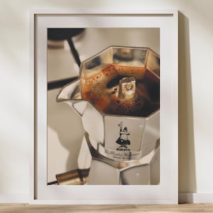 May include: A framed photograph of a silver stovetop espresso maker, filled with dark coffee. The coffee maker is in the centre of the frame, with the top open, revealing the rich, dark brew. The image is set against a neutral background.