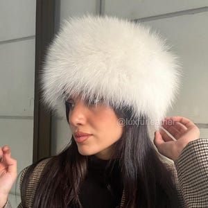 Op de afbeelding: Een pluizige, witte bont hoofdband. De hoofdband is het middelpunt, met een dichte, getextureerde uitstraling. Het model draagt een zwarte coltrui en een bruin-zwarte blazer met patroon. De Instagram-handle @luxfurleather is zichtbaar.