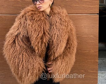 Puede incluir: Un abrigo de piel marrón y peludo. El abrigo tiene un tono cálido y terroso y tiene una apariencia voluminosa y texturizada. La imagen también muestra un vestido negro, botas negras y gafas de sol. El texto "@luxfurleather" es visible.