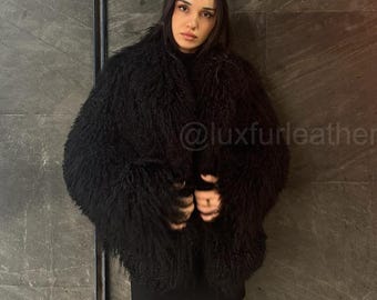 Puede incluir: Una mujer lleva una chaqueta negra peluda con un jersey de cuello alto negro y medias negras. La chaqueta tiene una textura esponjosa y se lleva abierta. La mujer está de pie frente a una pared gris texturizada. El texto "@luxfurleather" es visible en la pared.