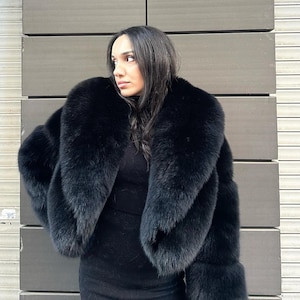May include: A black fur coat with a large collar. The coat is cropped and has long sleeves. The person wearing the coat is standing in front of a gray wall.