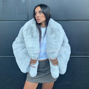 May include: A light blue faux fur coat with a wide collar. The coat is worn over a white top and a gray skirt. The text "@luxfurleather" is visible on the coat.