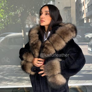 May include: A black fur coat with brown fur trim around the hood and cuffs. The coat is worn by a person standing outside. The Instagram handle @luxfurleather is visible on the coat.