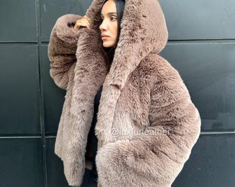 May include: A brown faux fur coat with a hood. The coat has a plush, textured appearance and a relaxed fit. The sleeves are long and voluminous. The coat is worn over a black top. The background is a dark gray wall.