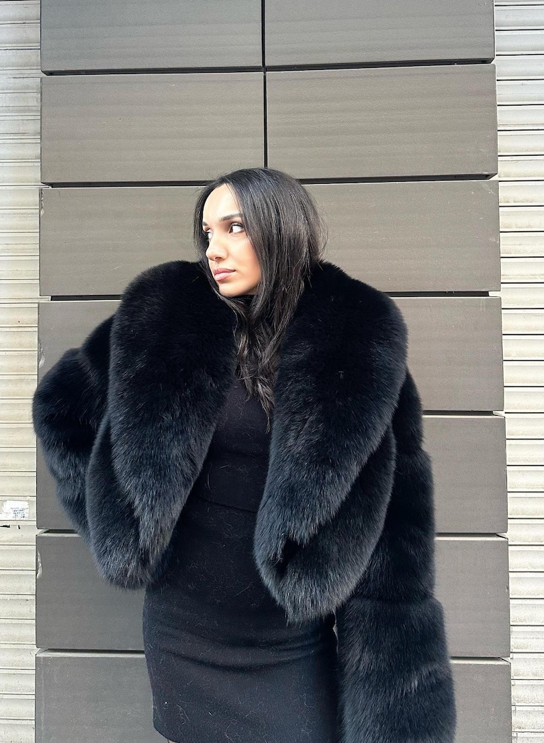 May include: A black fur coat with a large collar. The coat is cropped and has long sleeves. The person wearing the coat is standing in front of a gray wall.