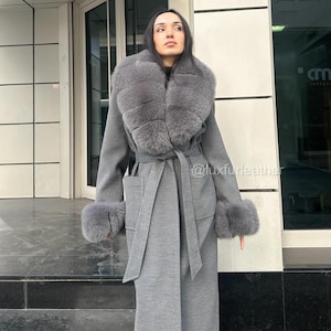 May include: A grey wool coat with a large grey fox fur collar and matching cuffs. The coat has a belt and a single pocket on the front.