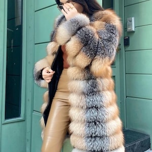 May include: A long, multi-toned fur coat with shades of brown, tan, and gray. The coat has a black lining and is worn with tan leather pants. The fur appears to be layered in horizontal bands. The coat is knee-length.