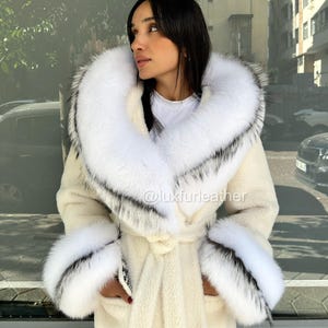May include: Cream-colored coat with a large white fur collar and cuffs, accented with black and gray details. The coat features a belted waist and is worn by a person. The Instagram handle @luxfurleather is visible.