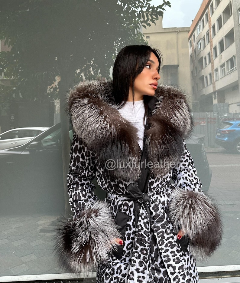 Op de afbeelding: Een lange jas met een zwart-wit luipaardprint, met een grote bontkraag en manchetten. De jas is in de taille vastgebonden met een zwarte riem. De tekst "@luxfurleather" is zichtbaar op de jas.