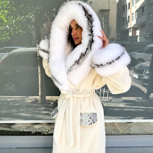 May include: Cream-coloured long coat with a large, fluffy white hood trimmed with black and grey fur. The coat features matching fur cuffs and pockets with a snakeskin pattern. A matching belt cinches the waist.