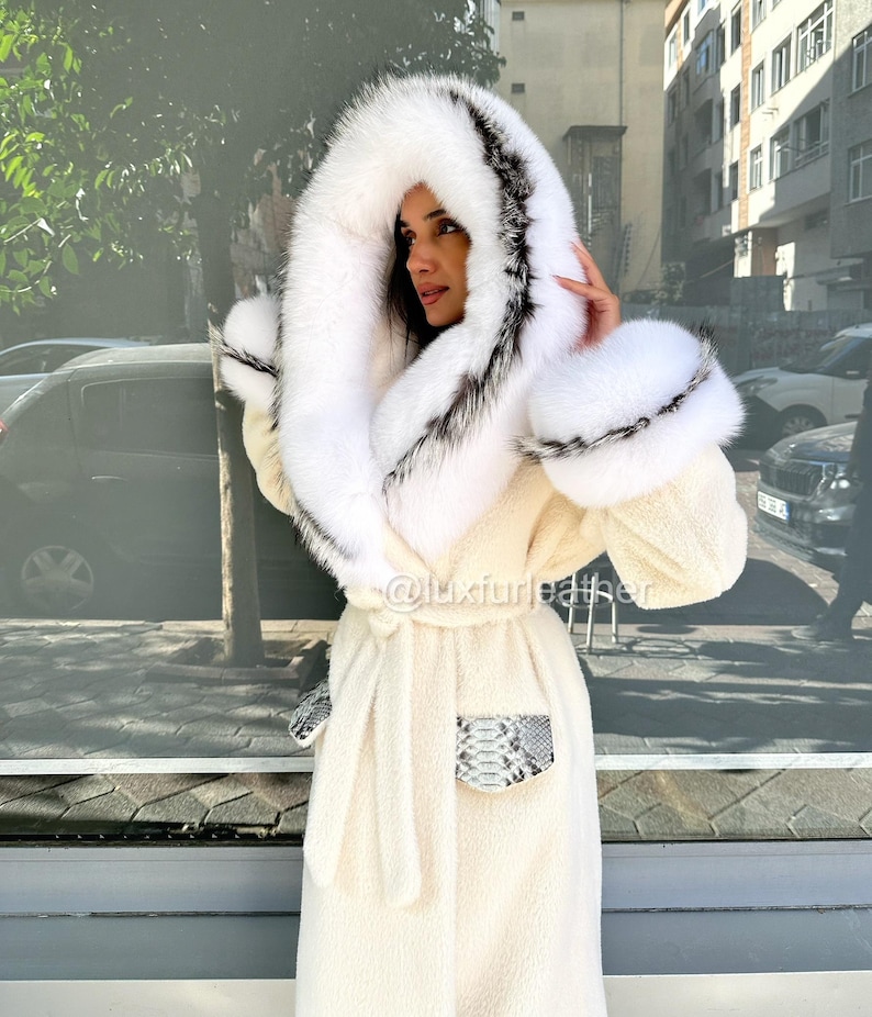 May include: Cream-colored long coat with a large, fluffy white hood trimmed with black and gray fur. The coat features matching fur cuffs and pockets with a snakeskin pattern. A matching belt cinches the waist.