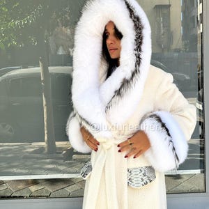 May include: Cream-colored long coat with a large hood and cuffs trimmed with white and black fur. The coat features a matching belt and pockets with a snakeskin pattern. The image includes the text "@luxfurleather".