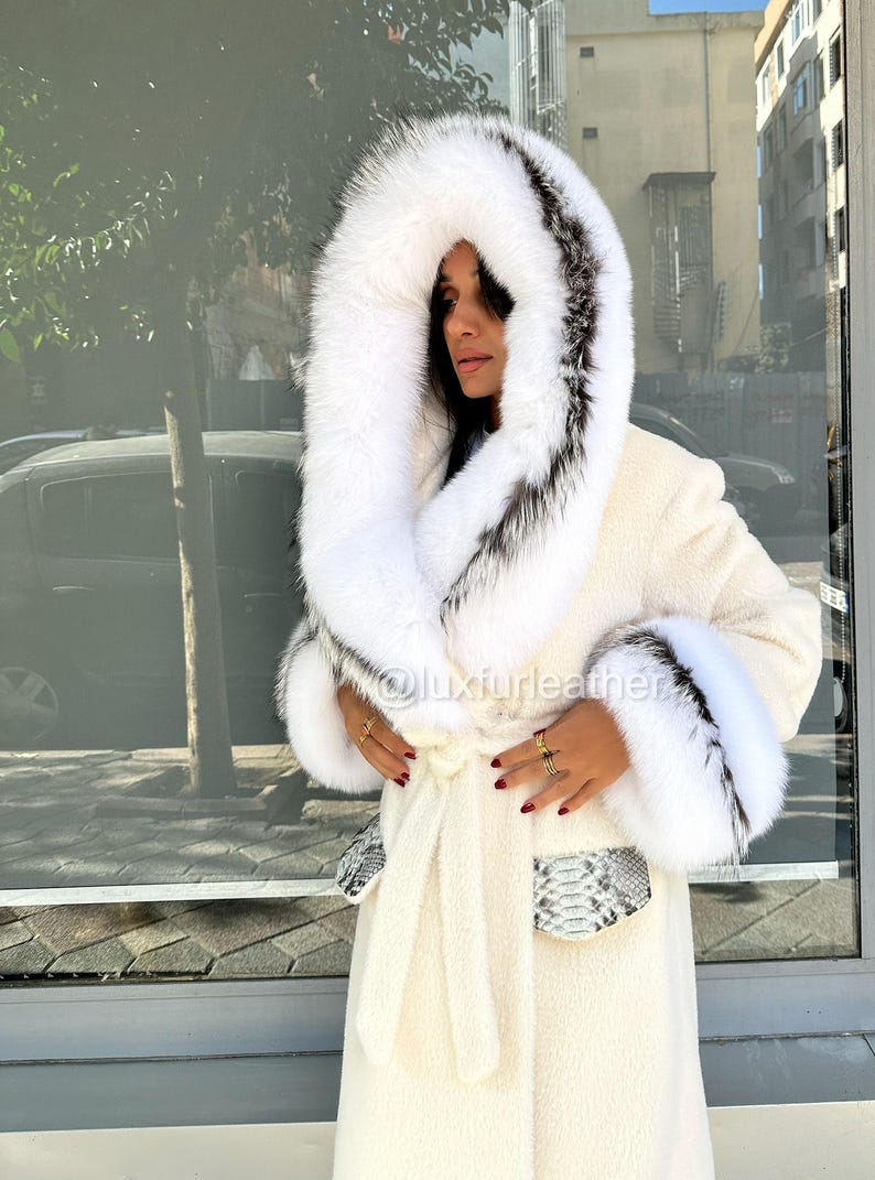 May include: Cream-colored long coat with a large hood and cuffs trimmed with white and black fur. The coat features a matching belt and pockets with a snakeskin pattern. The image includes the text "@luxfurleather".