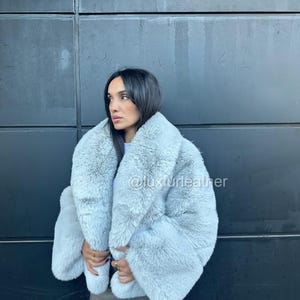 May include: A light blue faux fur coat with a wide collar and long sleeves. The coat is worn over a gray mini skirt. The text "@luxurleather" is visible on the coat. The coat appears soft and fluffy.