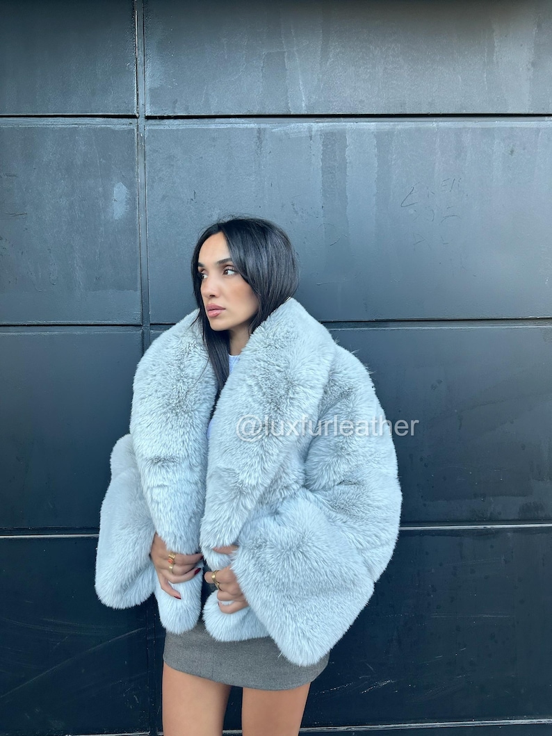 May include: A light blue faux fur coat with a wide collar and long sleeves. The coat is worn over a gray mini skirt. The text "@luxurleather" is visible on the coat. The coat appears soft and fluffy.